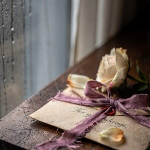 Lettre ancienne attachée avec un ruban et une rose fanée posée sur une table en bois près d’une fenêtre humide.