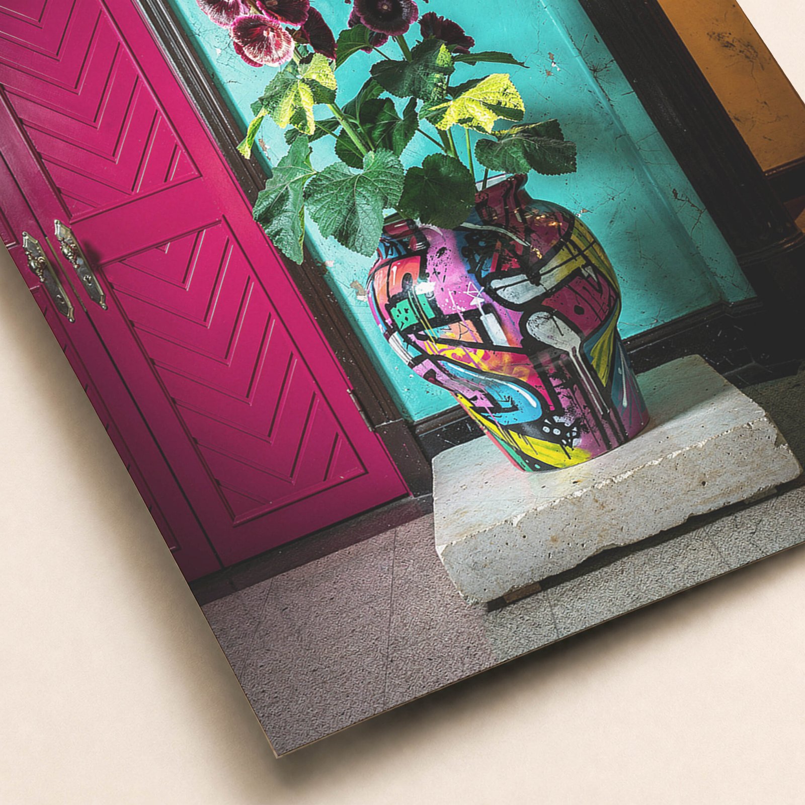 Gros plan sur une affiche avec vase coloré, fleurs pourpres, mur turquoise patiné et porte rose intense.